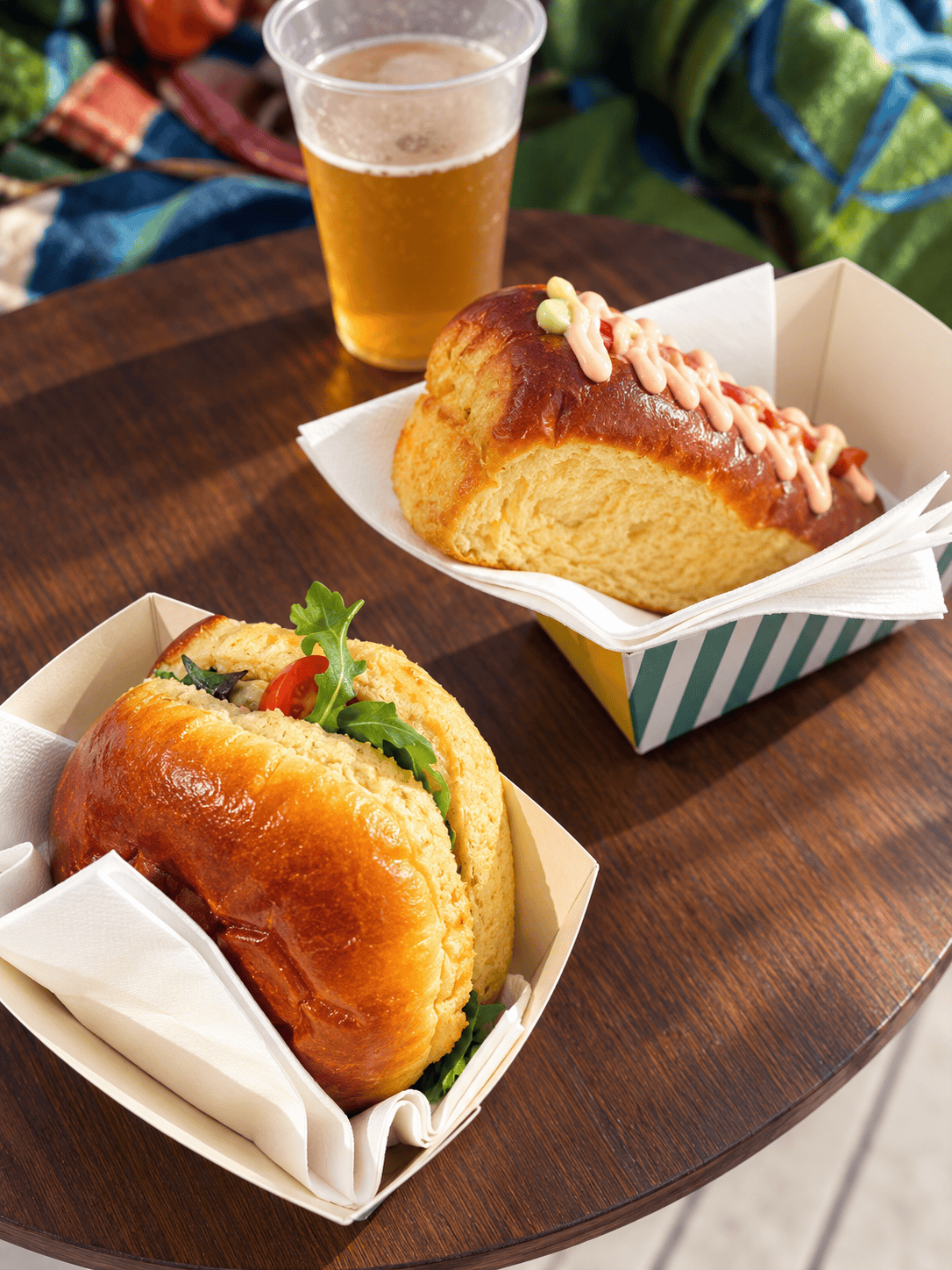 Two gourmet hot dog buns, one with greens and peppers, the other topped with sauce, sit on a wooden table with a cup of beer and a colorful blanket in the background.