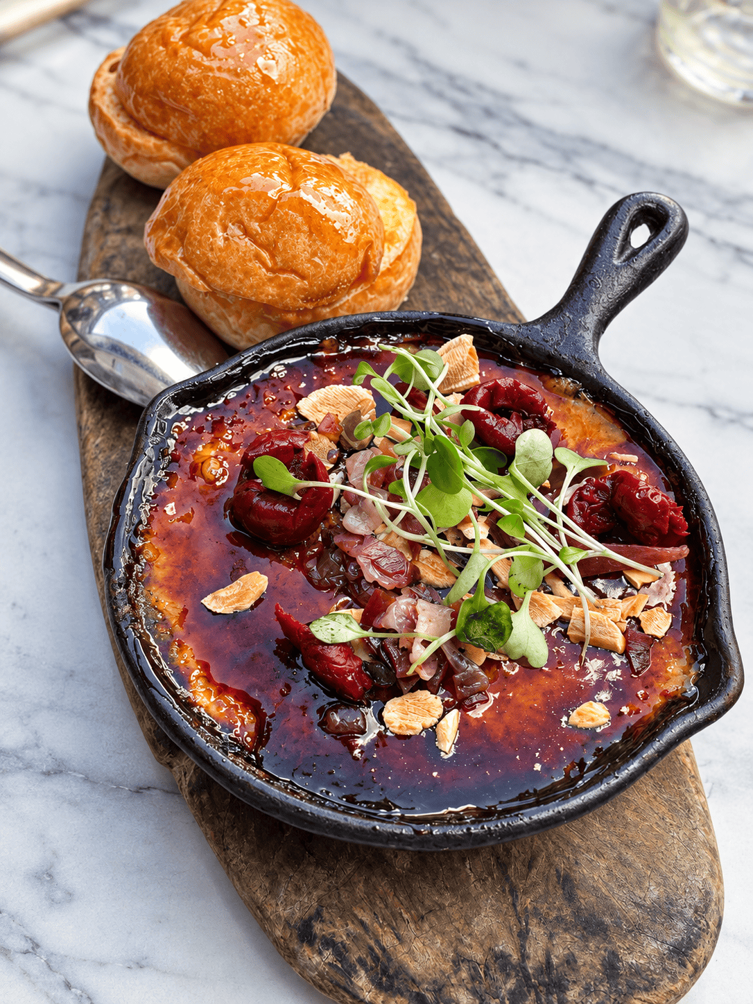 Crème brûlée topped with cherries, nuts, and microgreens in a small cast-iron skillet, served with two brioche rolls on a wooden board.